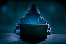 A hooded figure with his face covered in darkness hunched over a laptop with a light glow illuminating their silhouette. 
